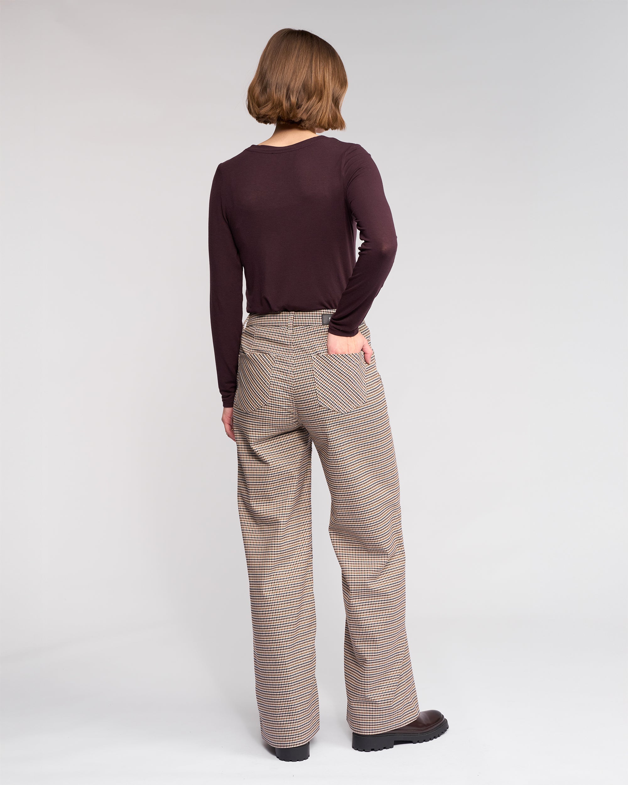 A person with chin-length brown hair faces away, wearing a long-sleeved dark brown top, Roxie Pant - Paige by Knewe Label, and dark shoes, with one hand in their back pocket against a plain background.