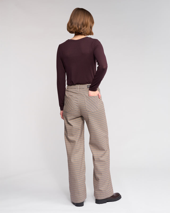 A person with chin-length brown hair faces away, wearing a long-sleeved dark brown top, Roxie Pant - Paige by Knewe Label, and dark shoes, with one hand in their back pocket against a plain background.