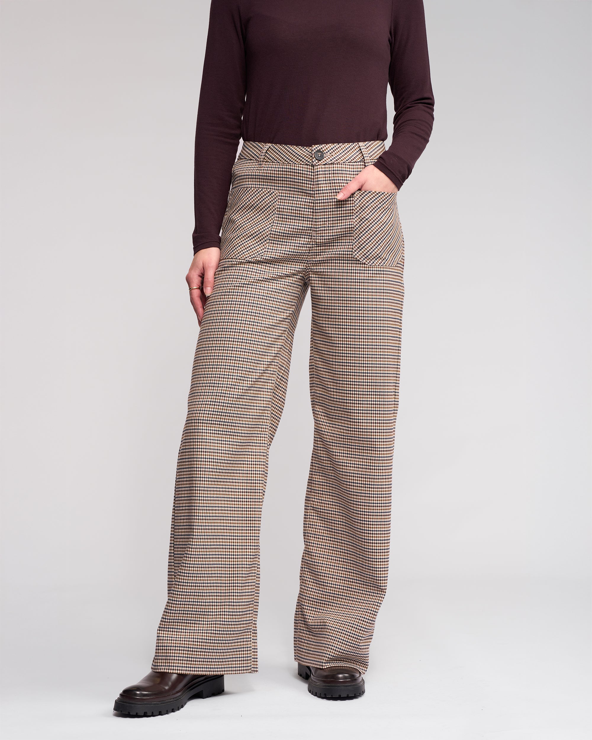 Wearing the Knewe Label Roxie Pant - Paige, high-waisted with a checked pattern and front pockets, the person stands against a plain background with one hand in a pocket. Only their body is visible; their face is not shown.