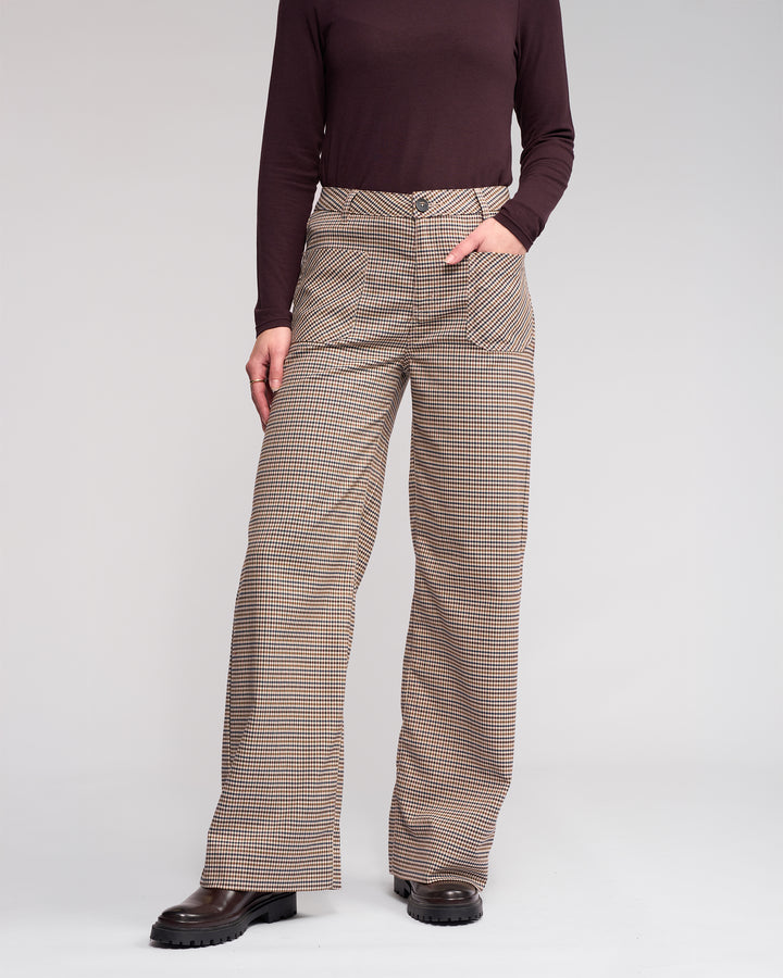 Wearing the Knewe Label Roxie Pant - Paige, high-waisted with a checked pattern and front pockets, the person stands against a plain background with one hand in a pocket. Only their body is visible; their face is not shown.