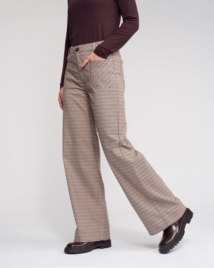 Wearing the Roxie Pant by Knewe Label, a person in a maroon long-sleeve top stands with one hand in their pocket. The brown wide-leg pants feature a subtle checked pattern and are paired with dark brown chunky boots against a plain light background.