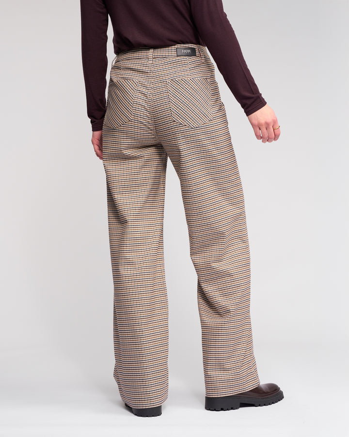 Person standing against a plain background, shown from the back wearing Knewe Label's Roxie Pant - Paige, featuring a brown checked wide-leg style with visible back pockets, paired with a dark long-sleeve top.