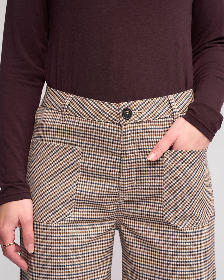 Someone wearing the Knewe Label Roxie Pant - Paige, featuring a high-waisted wide-leg checked design with large front pockets, paired with a long-sleeved dark brown top, stands with one hand in a pocket and the other at their side.