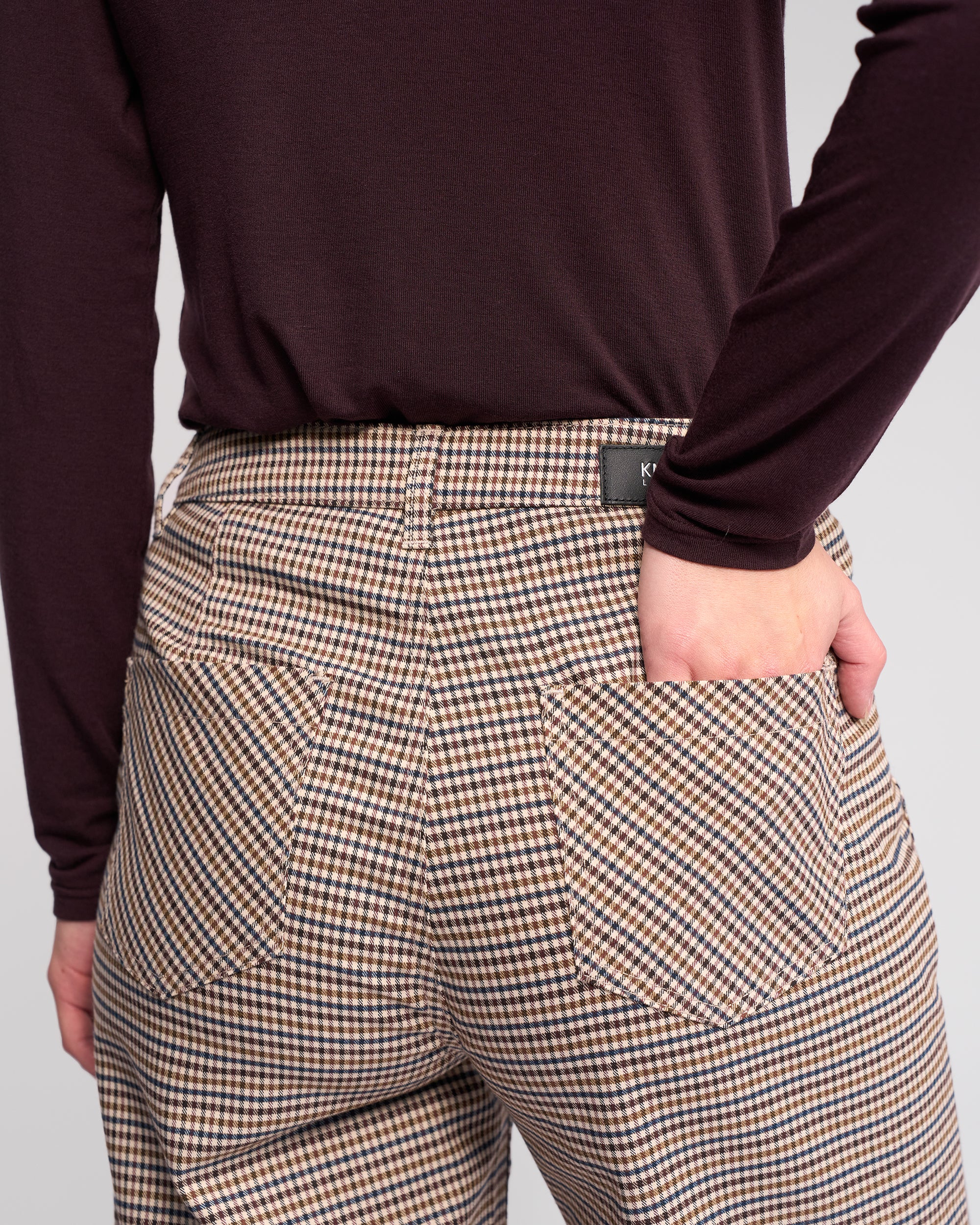 A person wears the Roxie Pant by Knewe Label—high-waisted, wide-leg checked pants with large back pockets. Facing away from the camera, their hand is partly in a back pocket, paired with a dark long-sleeved top.