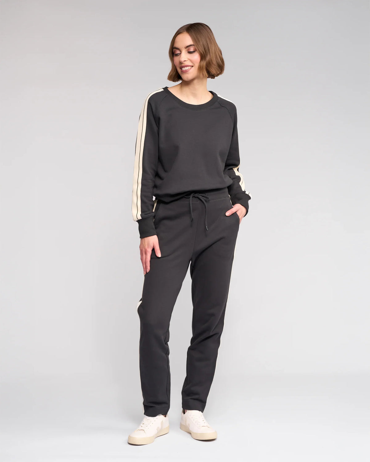 A woman stands smiling in Knewe Label's Rebel Pant - Slate, a matching black cotton set with white stripes and slim-leg ankle-grazer fit, paired with white sneakers, against a plain light gray background.