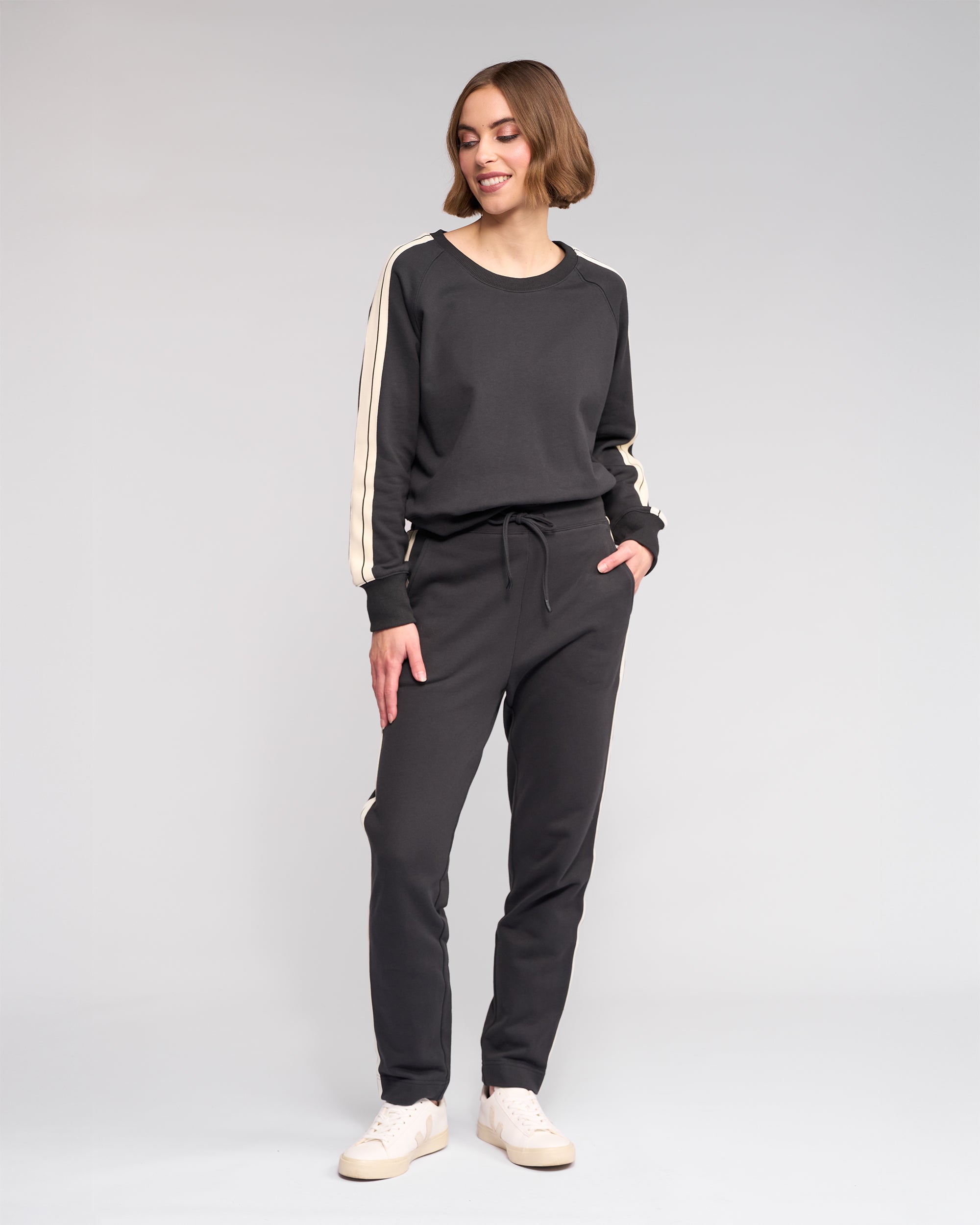 A woman stands smiling in Knewe Label's Rebel Pant - Slate, a matching black cotton set with white stripes and slim-leg ankle-grazer fit, paired with white sneakers, against a plain light gray background.