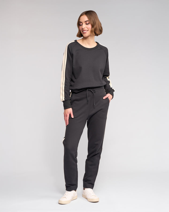 A woman stands smiling in Knewe Label's Rebel Pant - Slate, a matching black cotton set with white stripes and slim-leg ankle-grazer fit, paired with white sneakers, against a plain light gray background.