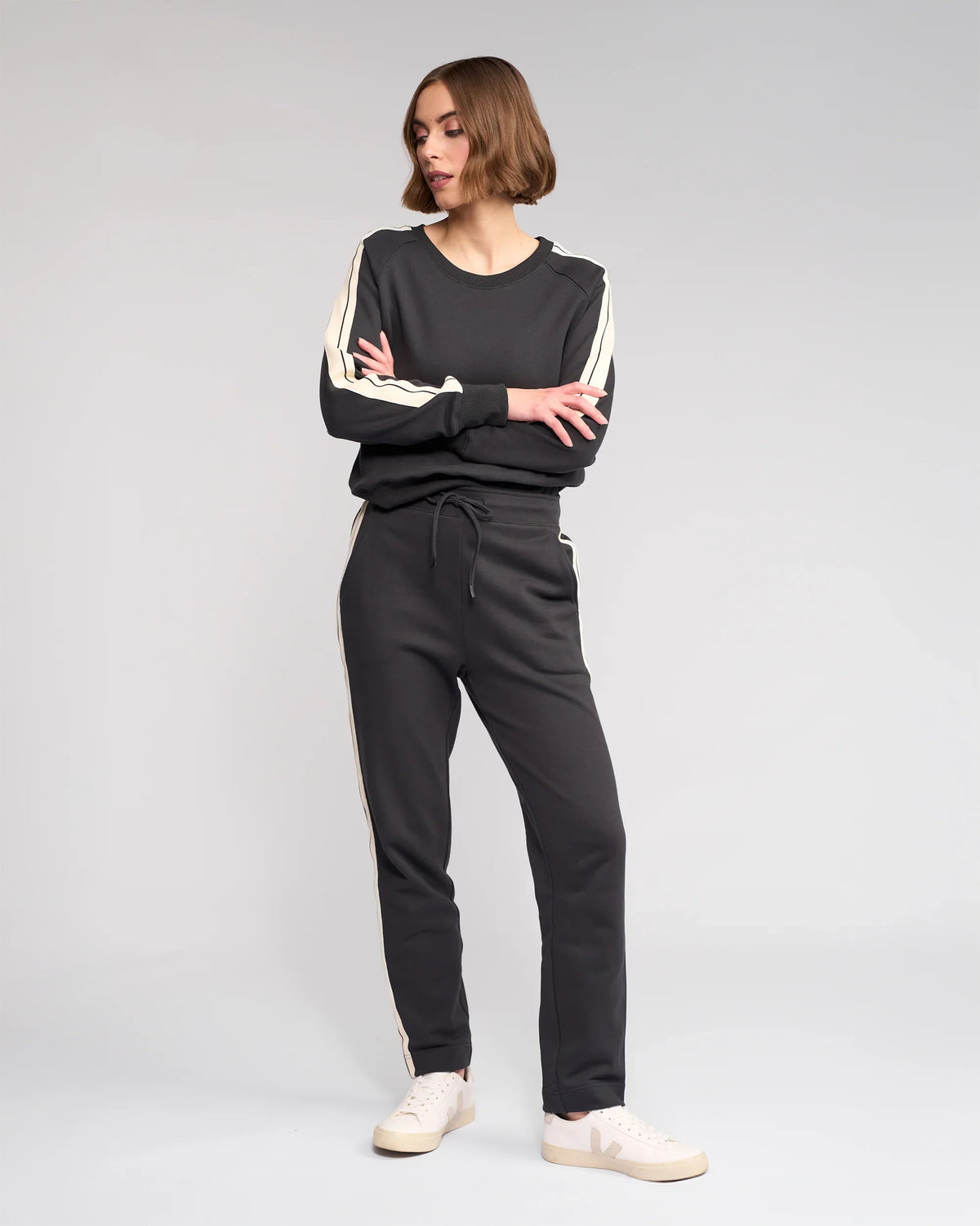 A woman poses with arms crossed against a plain backdrop, wearing Knewe Label’s Rebel Pant in Slate—slim-leg, ankle-grazer pants with white stripes—and white sneakers.
