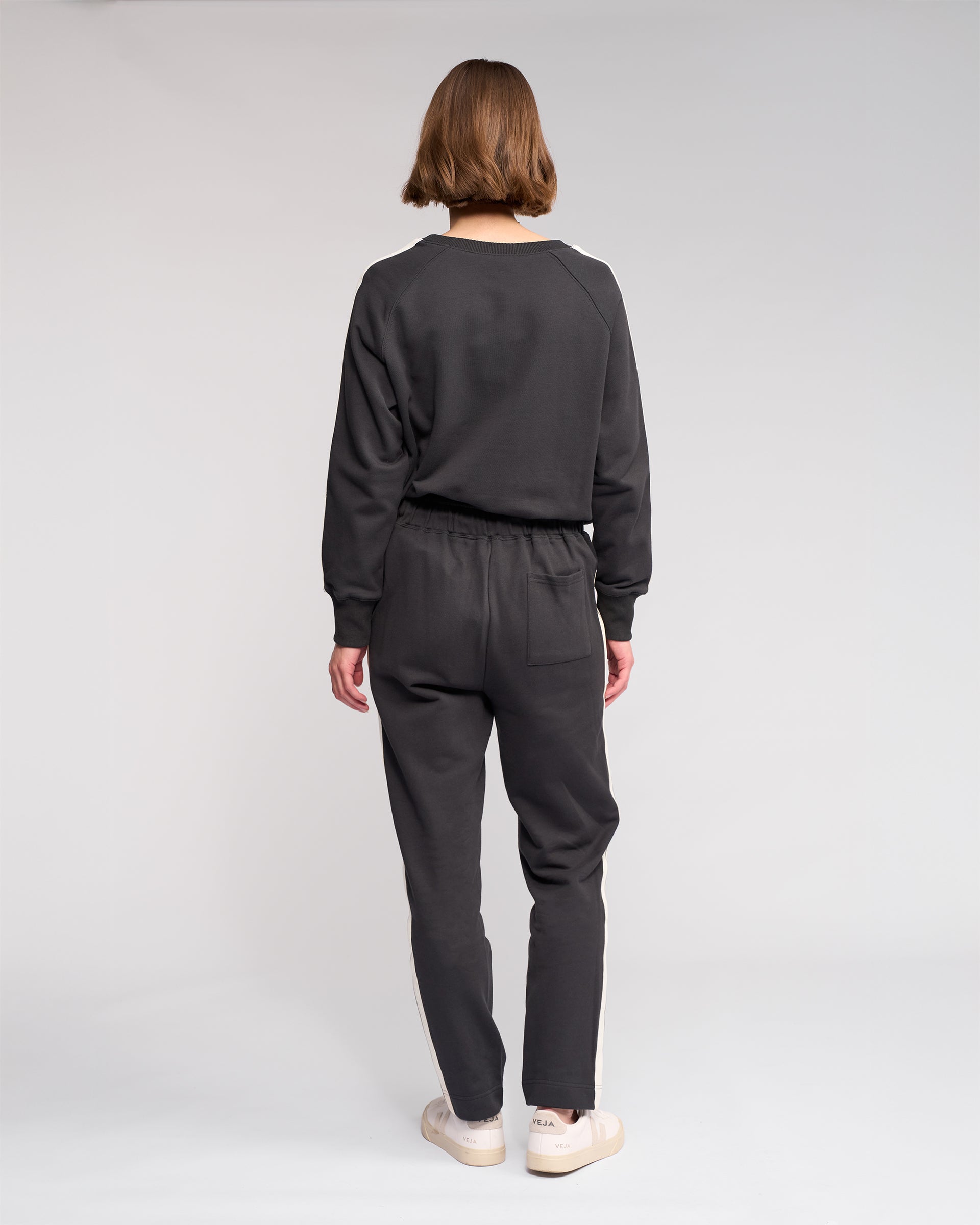 A person with short hair faces away from the camera, wearing Knewe Label’s Rebel Pant in Slate and a dark grey sweatshirt with white sneakers, standing against a plain light background.