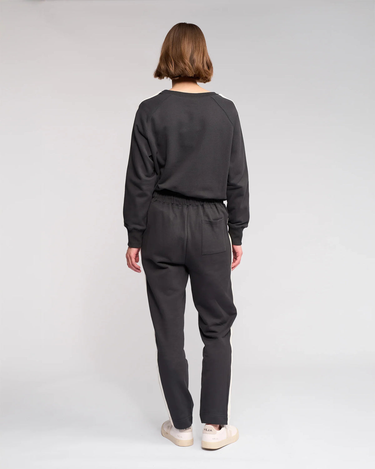 A person with short hair faces away from the camera, wearing Knewe Label’s Rebel Pant in Slate and a dark grey sweatshirt with white sneakers, standing against a plain light background.