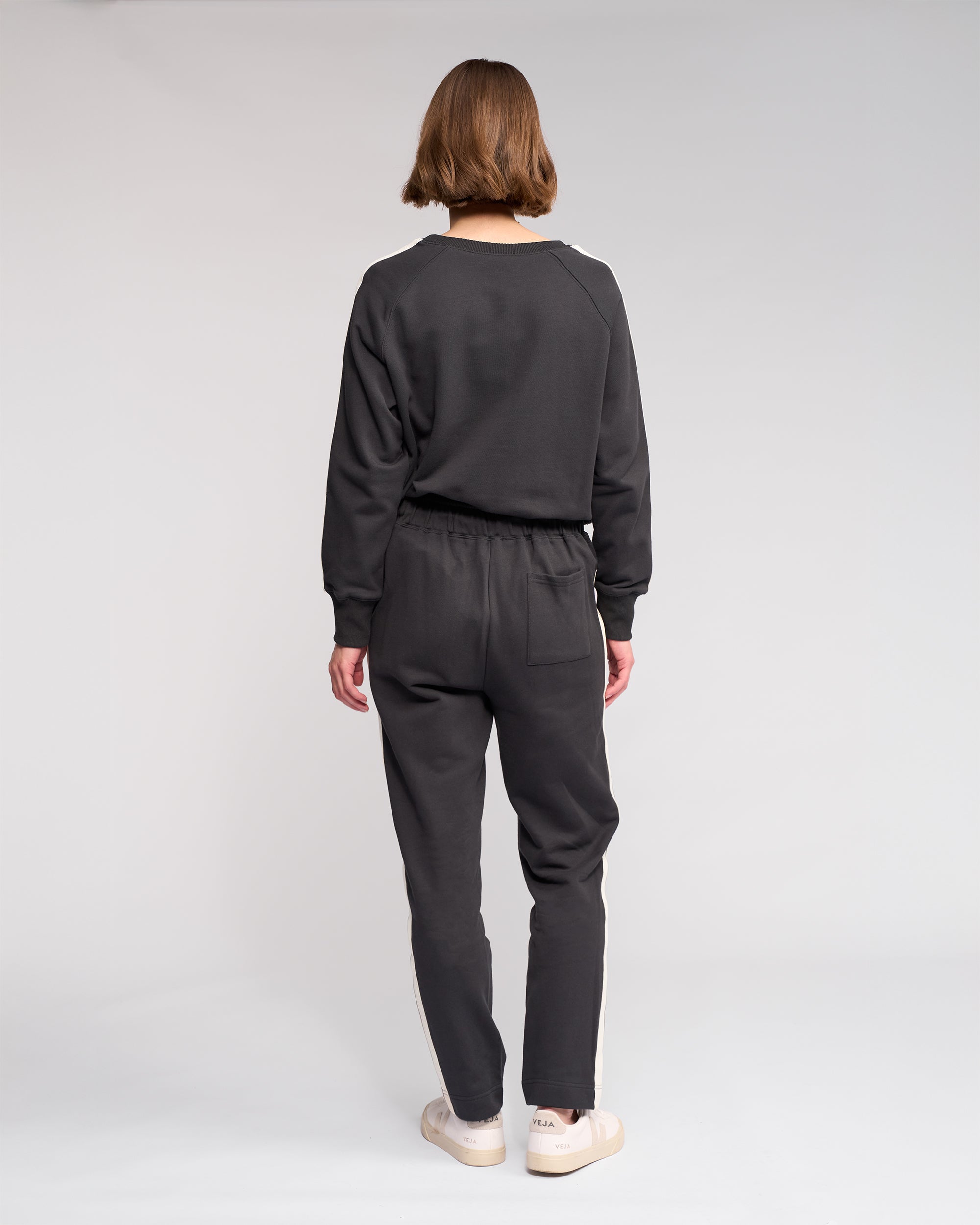 A person with short hair faces away from the camera, wearing Knewe Label’s Rebel Pant in Slate and a dark grey sweatshirt with white sneakers, standing against a plain light background.