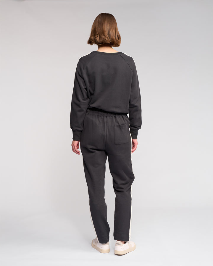 A person with short hair faces away from the camera, wearing Knewe Label’s Rebel Pant in Slate and a dark grey sweatshirt with white sneakers, standing against a plain light background.