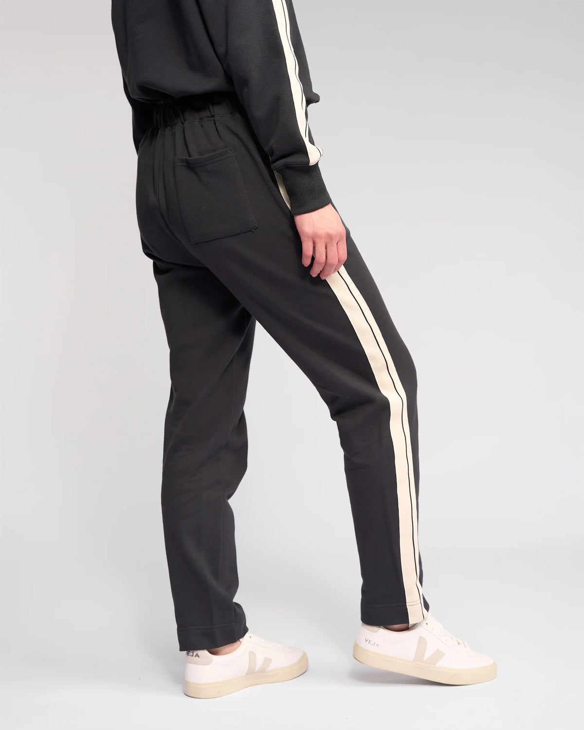A person wearing the Knewe Label Rebel Pant - Slate, dark grey slim-leg ankle-grazer joggers with white side stripes and white sneakers, stands with one leg forward, photographed from the back against a plain light background.