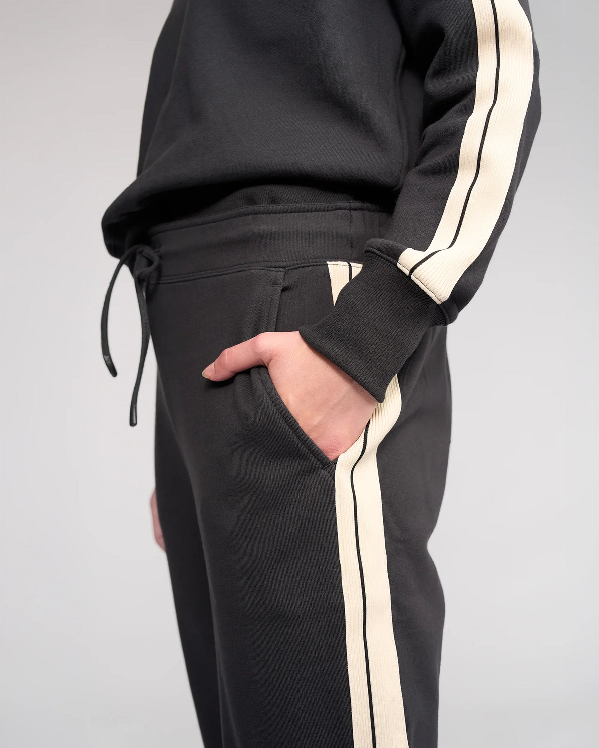 A person wears Knewe Label’s Rebel Pant in Slate—a black, slim-leg ankle-grazer with cream stripes and soft cotton fabric—shown from midsection to upper thigh, one hand in the pocket.