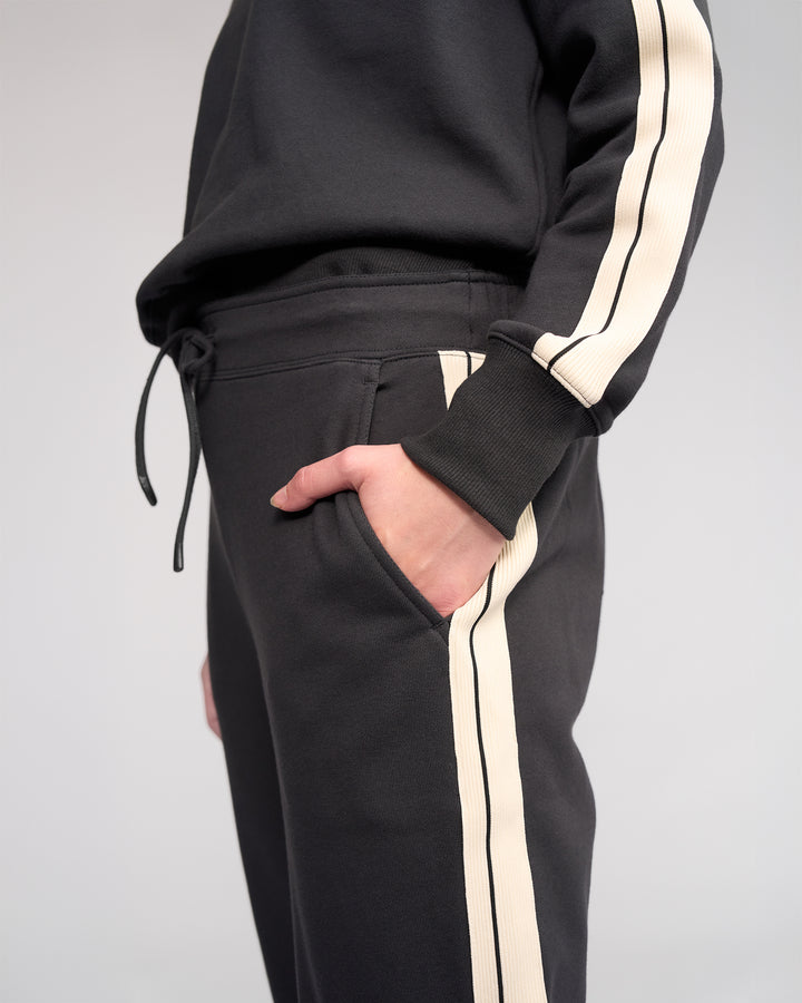A person wears Knewe Label’s Rebel Pant in Slate—a black, slim-leg ankle-grazer with cream stripes and soft cotton fabric—shown from midsection to upper thigh, one hand in the pocket.