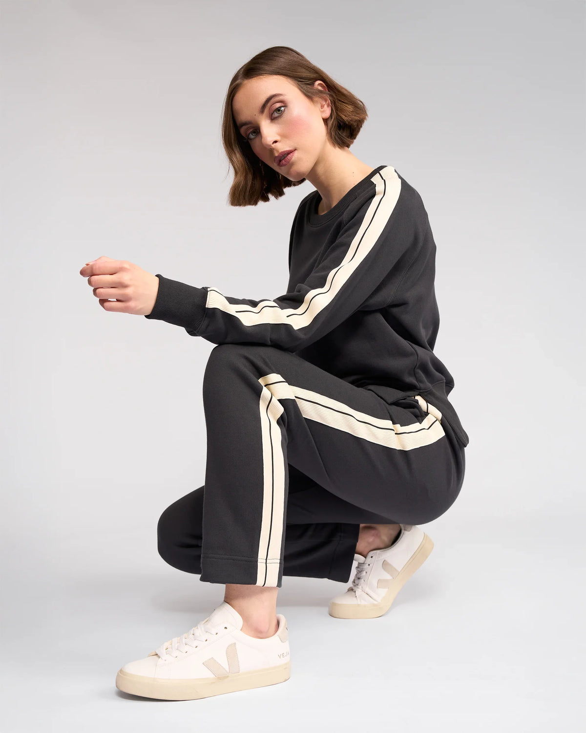 A woman with short brown hair squats sideways against a plain background, wearing Knewe Label’s Rebel Pant in Slate—a soft cotton, slim-leg ankle-grazer—and cream sneakers.