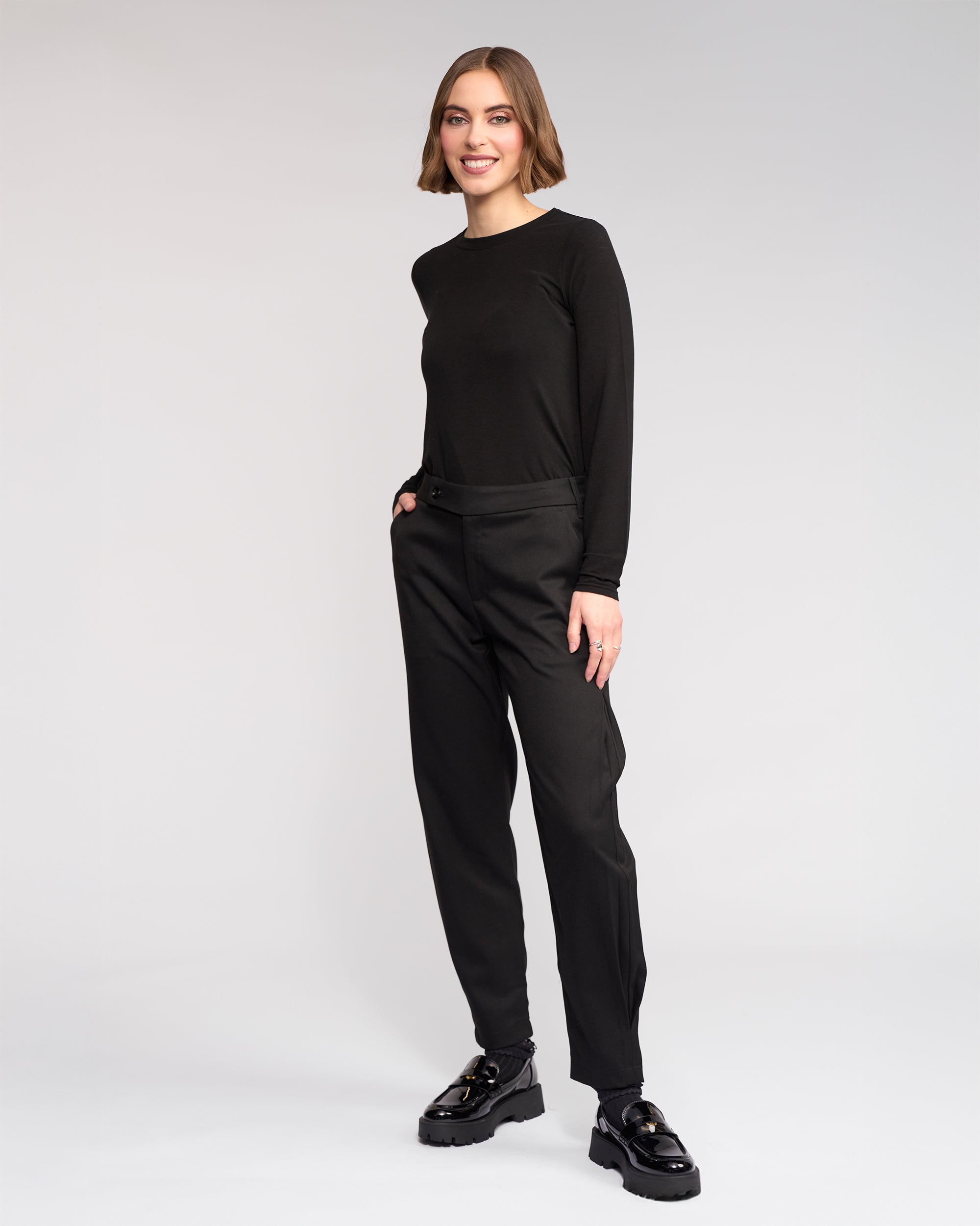 A woman with a bob haircut smiles, hands in pockets, wearing the Knewe Label Jude Pant in black with a long-sleeve black top and chunky shoes, standing against a plain light gray background.