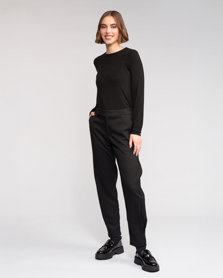 A woman with a bob haircut smiles, hands in pockets, wearing the Knewe Label Jude Pant in black with a long-sleeve black top and chunky shoes, standing against a plain light gray background.