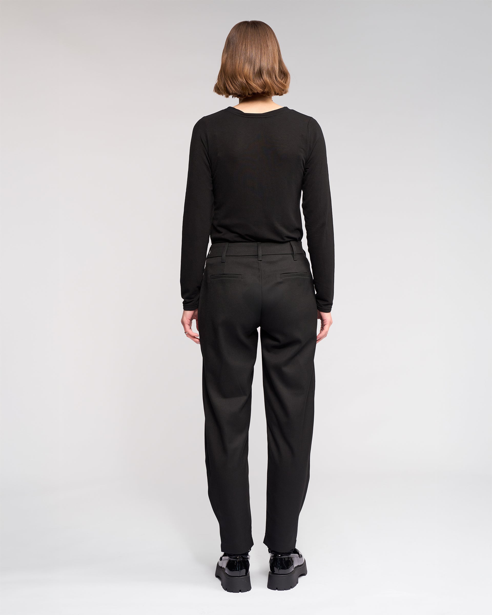 A person with short hair wears a black long-sleeve shirt, the Jude Pant - Black from Knewe Label, and black shoes, standing with their back to the camera against a plain white background.
