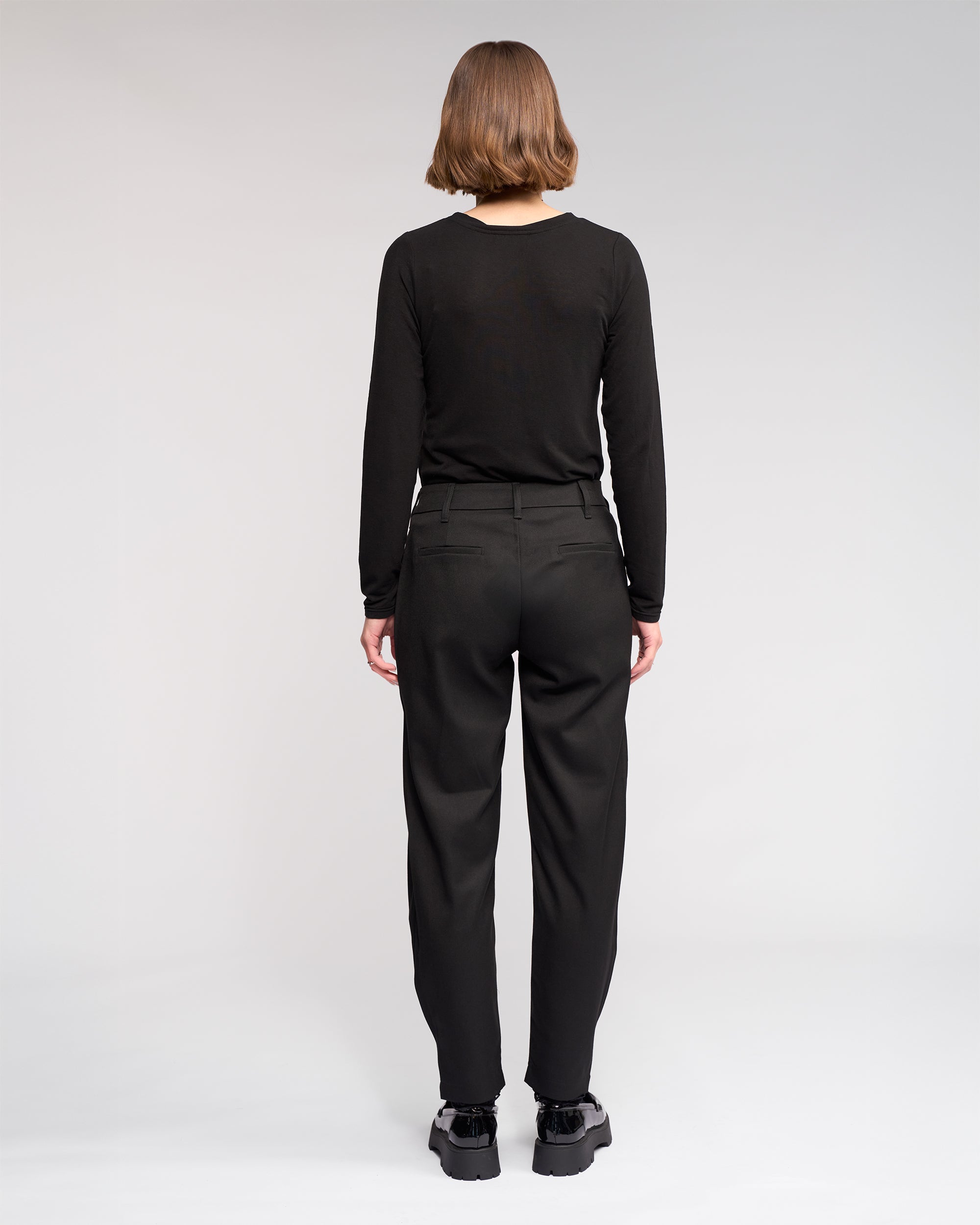 A person with short hair wears a black long-sleeve shirt, the Jude Pant - Black from Knewe Label, and black shoes, standing with their back to the camera against a plain white background.