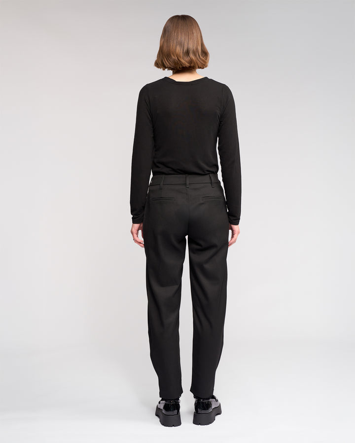 A person with short hair wears a black long-sleeve shirt, the Jude Pant - Black from Knewe Label, and black shoes, standing with their back to the camera against a plain white background.