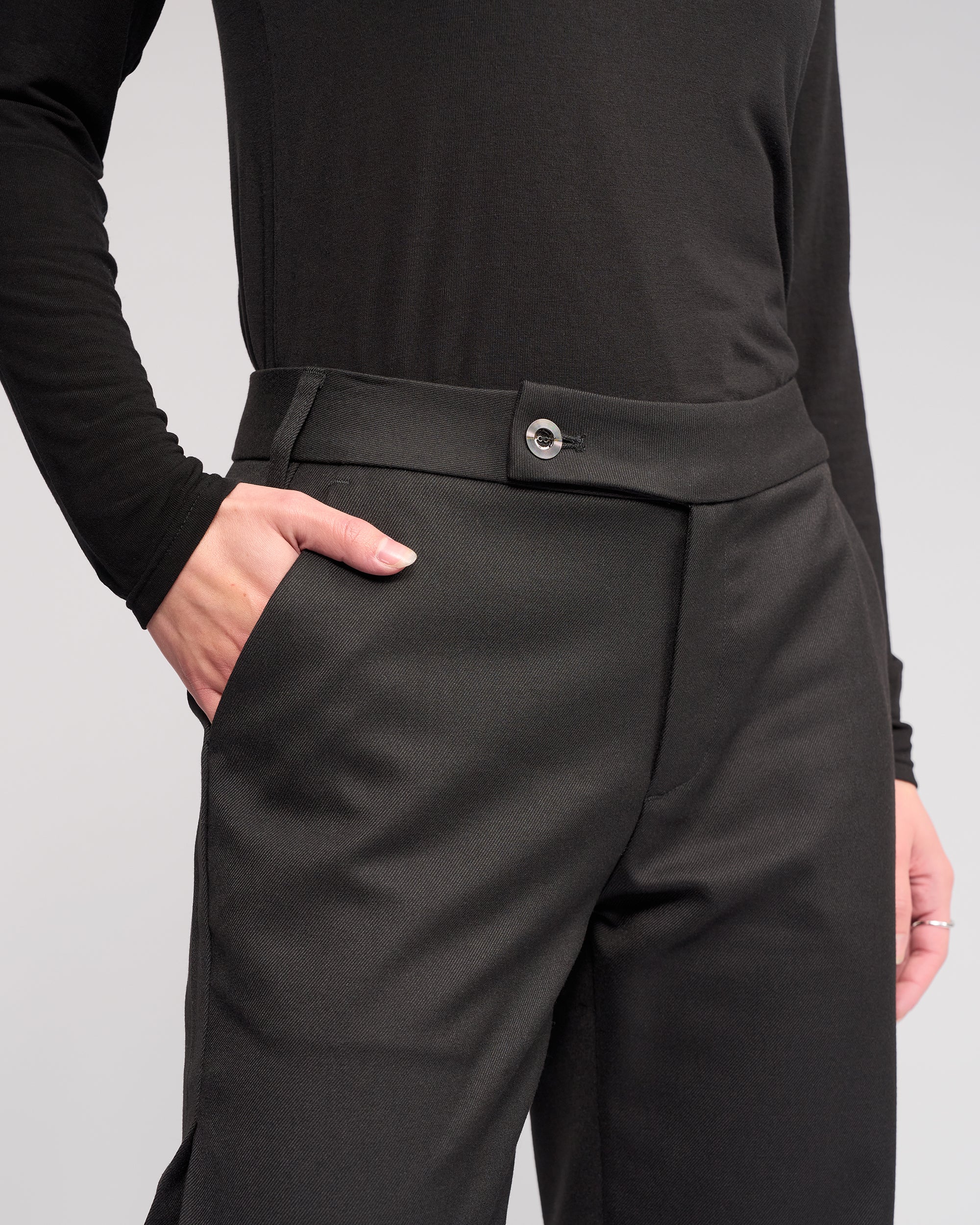 Model wears the Knewe Label Jude Pant in Black, styled with a relaxed leg. Shot from the waist down against a plain light background, their right hand is in their pocket.