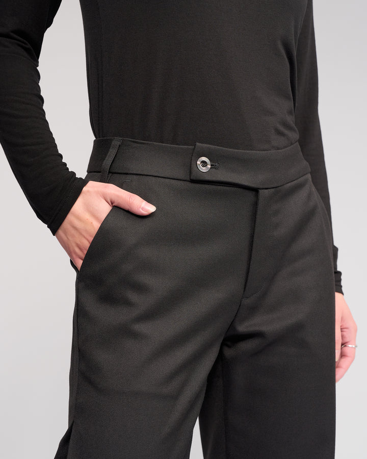 Model wears the Knewe Label Jude Pant in Black, styled with a relaxed leg. Shot from the waist down against a plain light background, their right hand is in their pocket.