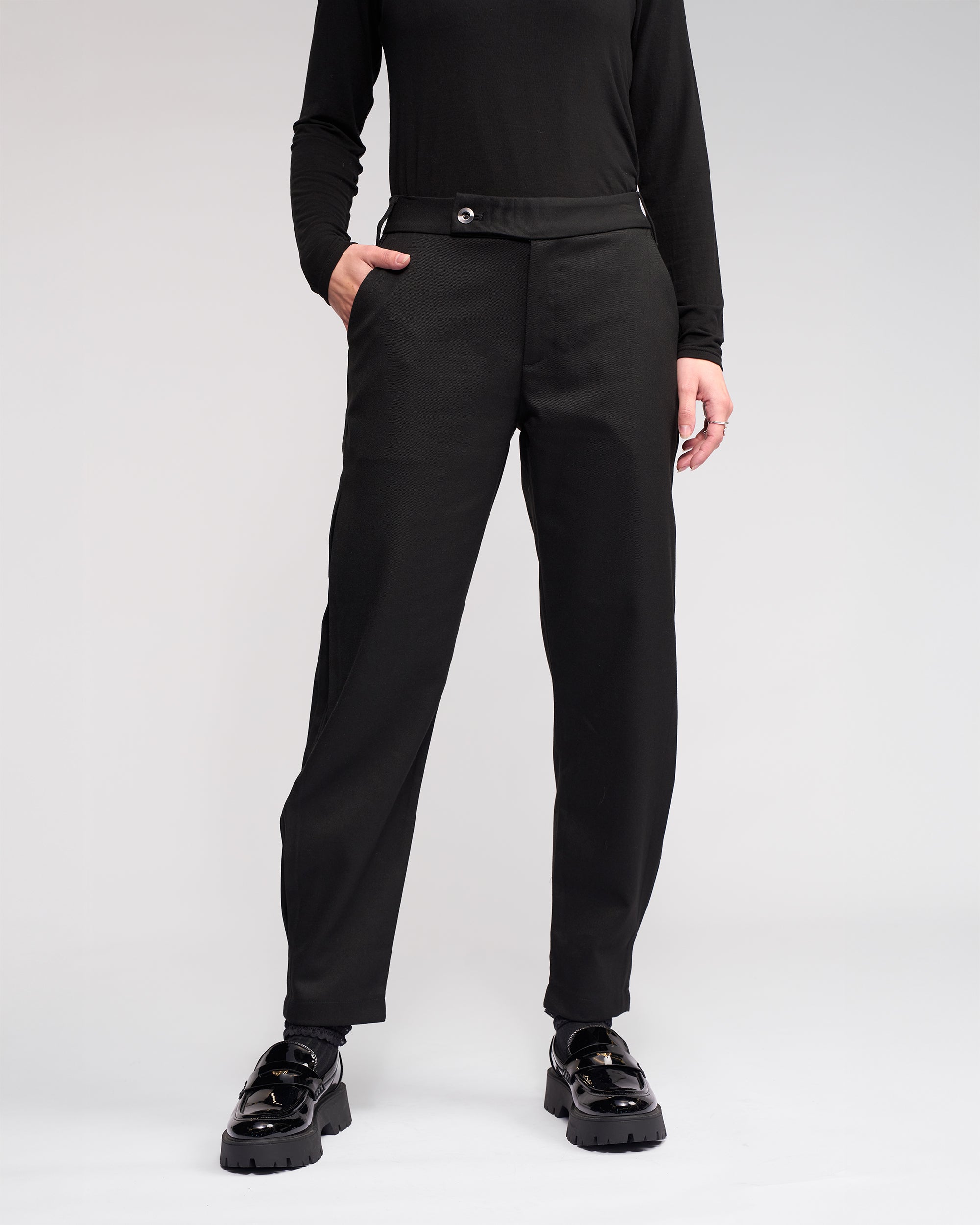 Wearing the Jude Pant - Black by Knewe Label, a person stands with one hand in their pocket against a plain light background. Their face is not visible.