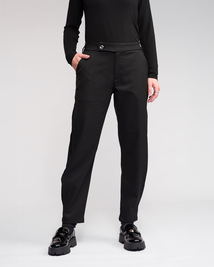 Wearing the Jude Pant - Black by Knewe Label, a person stands with one hand in their pocket against a plain light background. Their face is not visible.