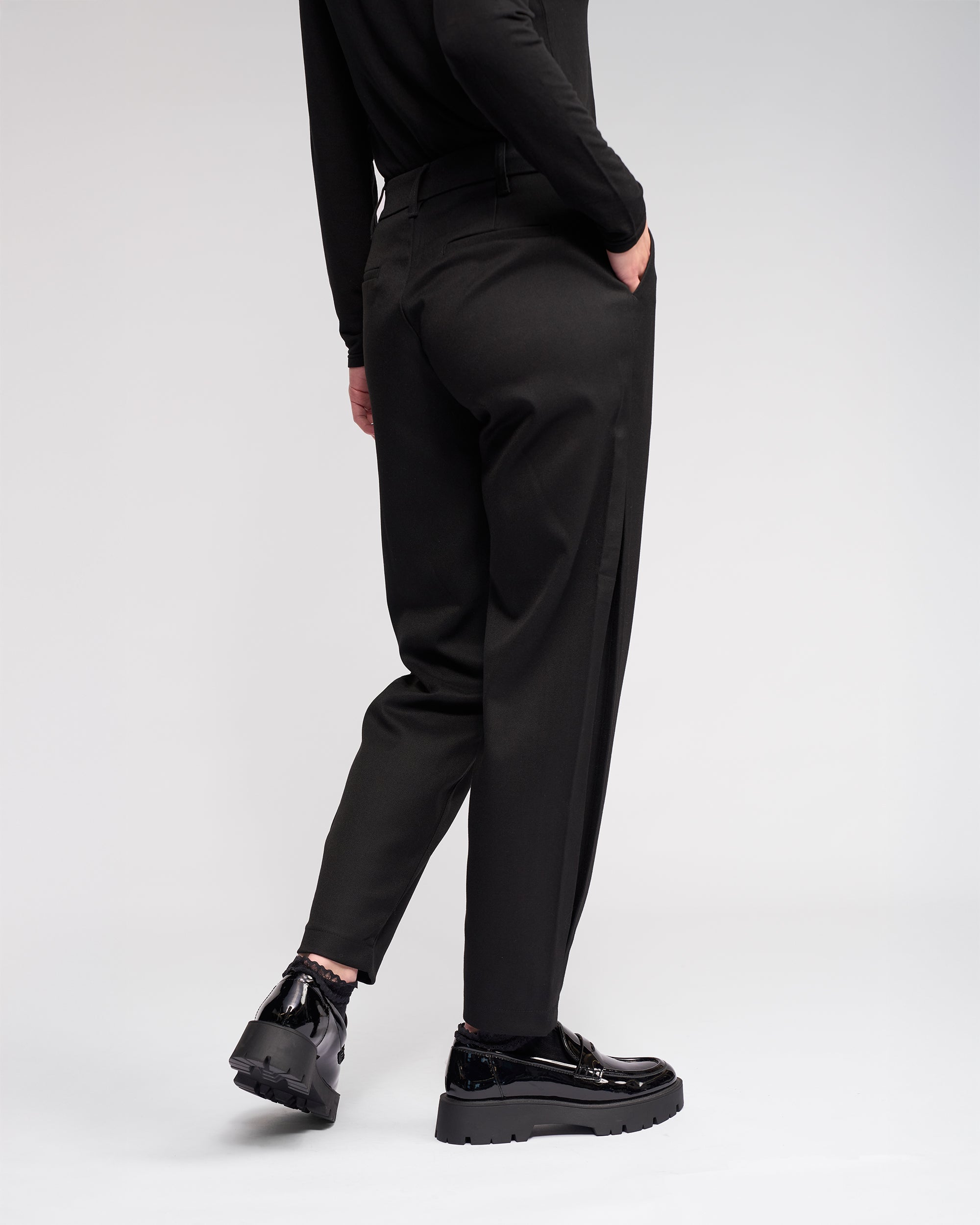 A person models the Knewe Label Jude Pant in black, styled with a matching black top and platform shoes, standing with one hand in their pocket and facing slightly away from the camera against a plain light background.