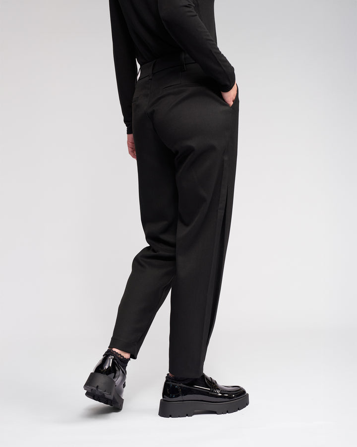 A person models the Knewe Label Jude Pant in black, styled with a matching black top and platform shoes, standing with one hand in their pocket and facing slightly away from the camera against a plain light background.