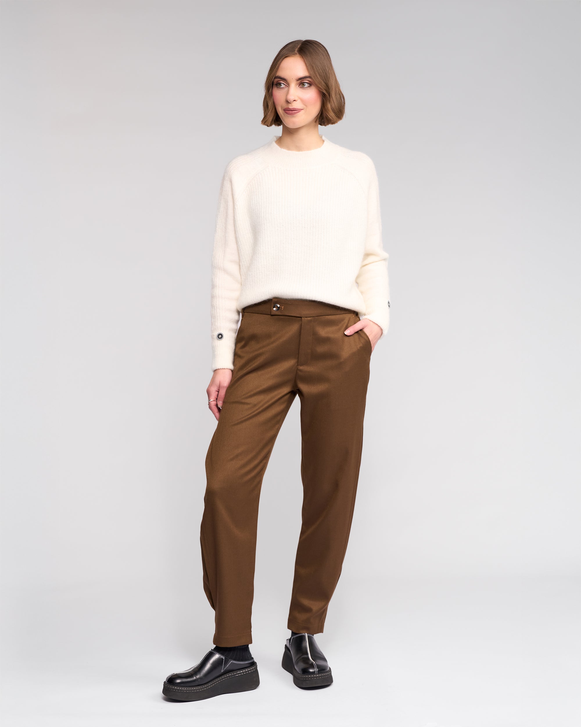 A woman with short brown hair smiles softly against a plain background, wearing the Knewe Label Jude Pant in Cedar, a cream-colored sweater, and black shoes, with one hand in her pocket.