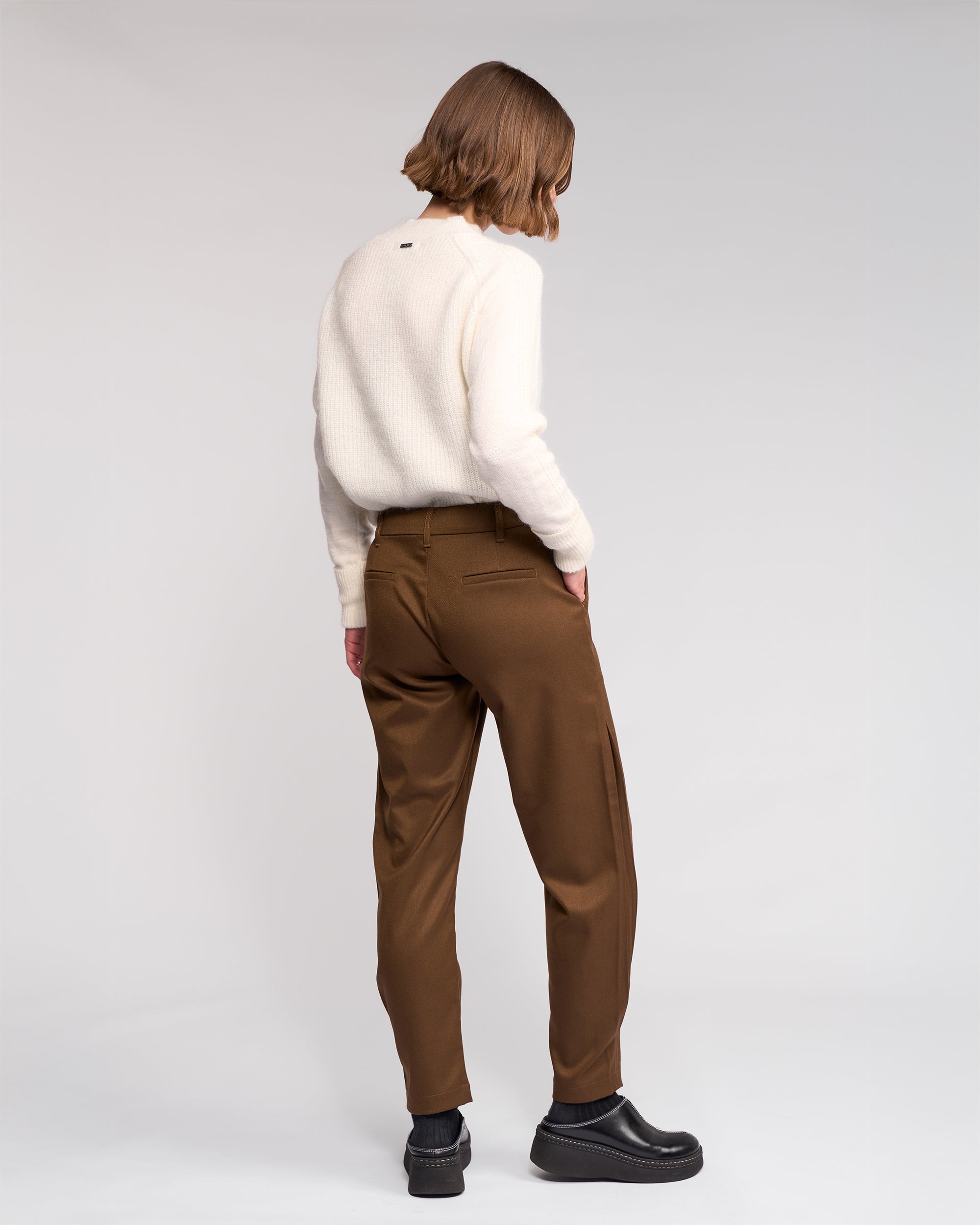 A person with short, light brown hair stands facing away, wearing a cream sweater and the Knewe Label Jude Pant in Cedar—brown relaxed-leg pants with a modern finish—paired with black shoes against a plain light background.