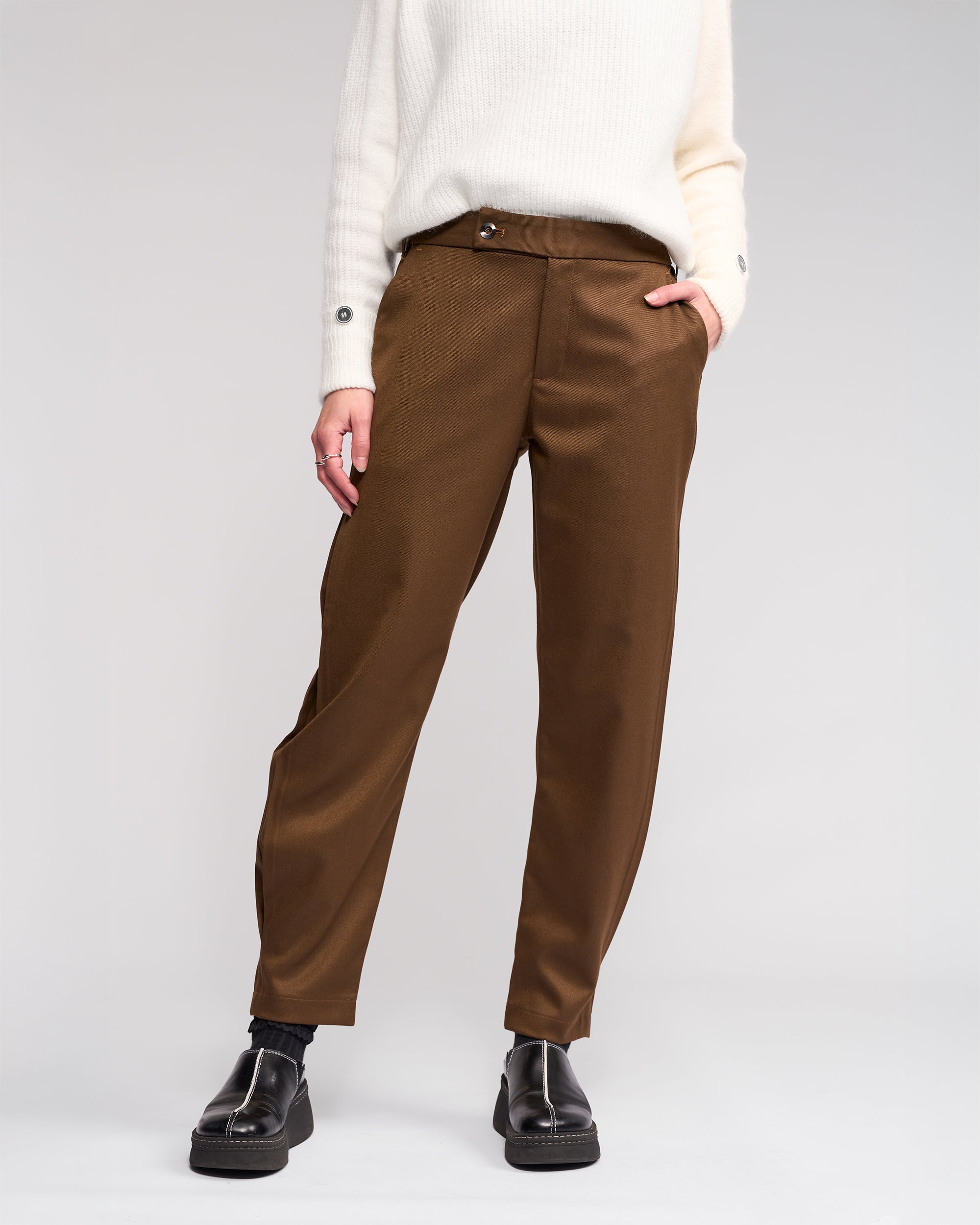 Wearing the Knewe Label Jude Pant in Cedar, styled with a white sweater and black shoes, hands in pockets. The face is not visible, keeping focus on the modern finish of this relaxed-leg ankle-grazer look against a plain light background.