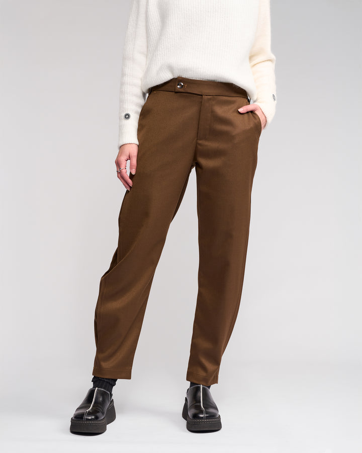 Wearing the Knewe Label Jude Pant in Cedar, styled with a white sweater and black shoes, hands in pockets. The face is not visible, keeping focus on the modern finish of this relaxed-leg ankle-grazer look against a plain light background.