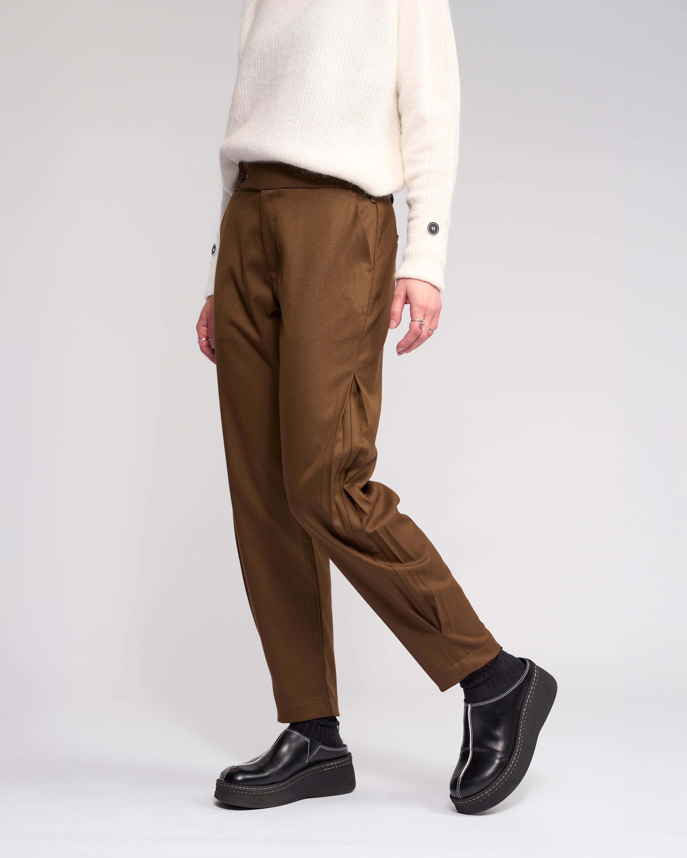 A person wears the Knewe Label Jude Pant in Cedar, paired with a white sweater and black platform shoes. Only their lower half is visible, highlighting the modern finish of these relaxed-leg, ankle-grazer pants against a plain light background.