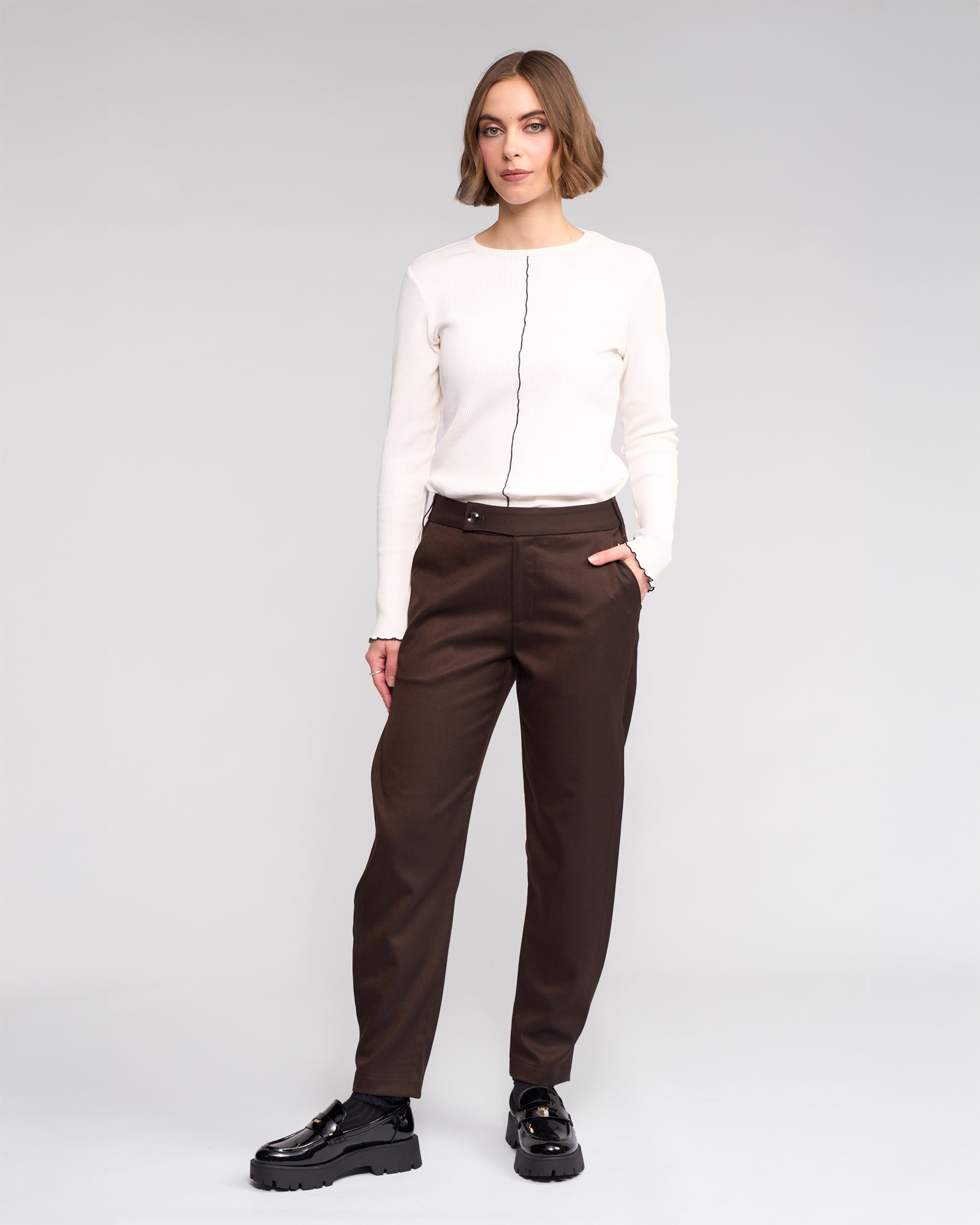 A person stands facing forward in a white long-sleeve top with a black vertical line, Knewe Label's Jude Pant in Chocolate, and black shiny shoes. One hand is in their pocket against a plain, light-colored background.