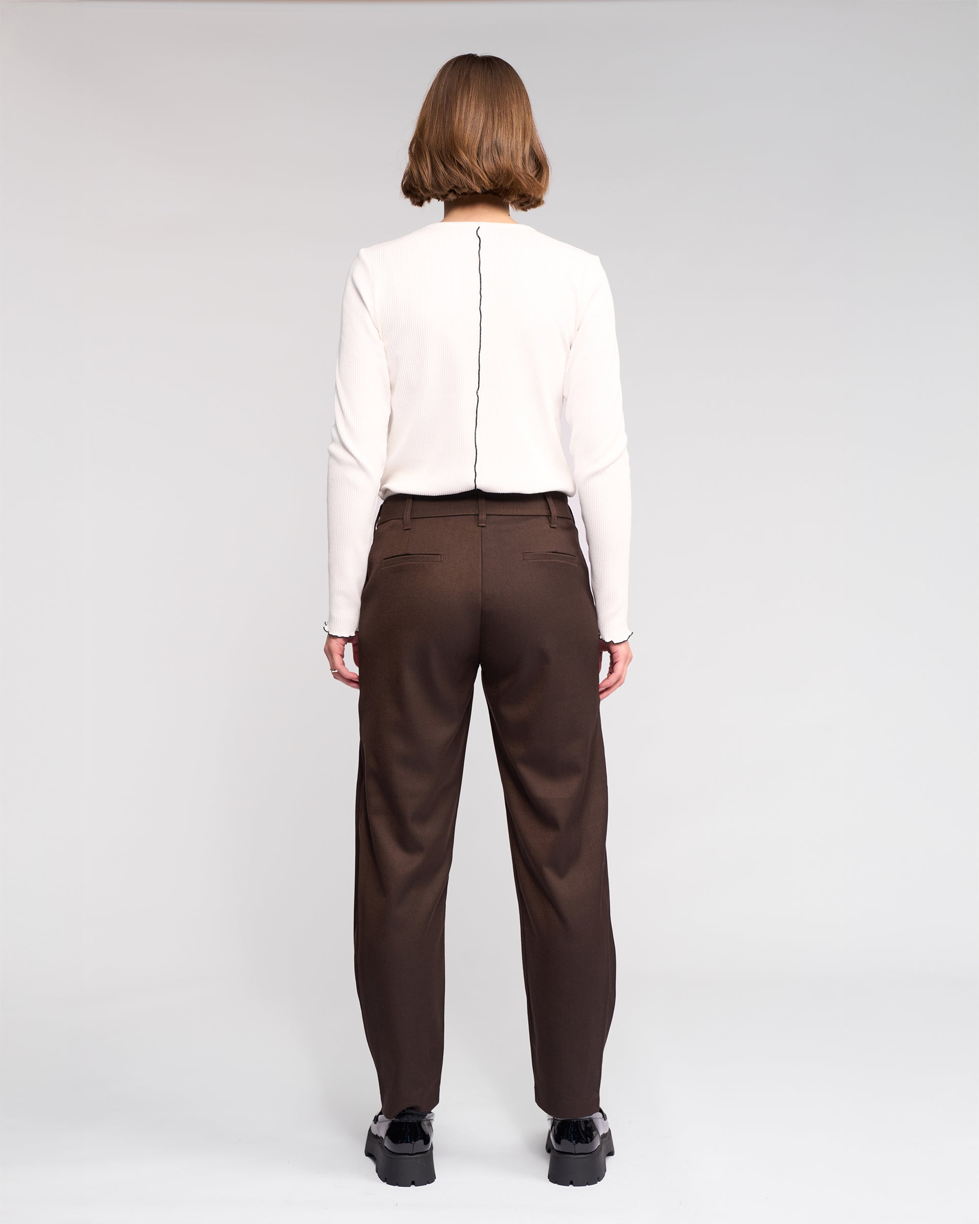 A person with short hair stands facing away against a plain light background, wearing a white long-sleeve top, black shoes, and the Jude Pant in Chocolate by Knewe Label.