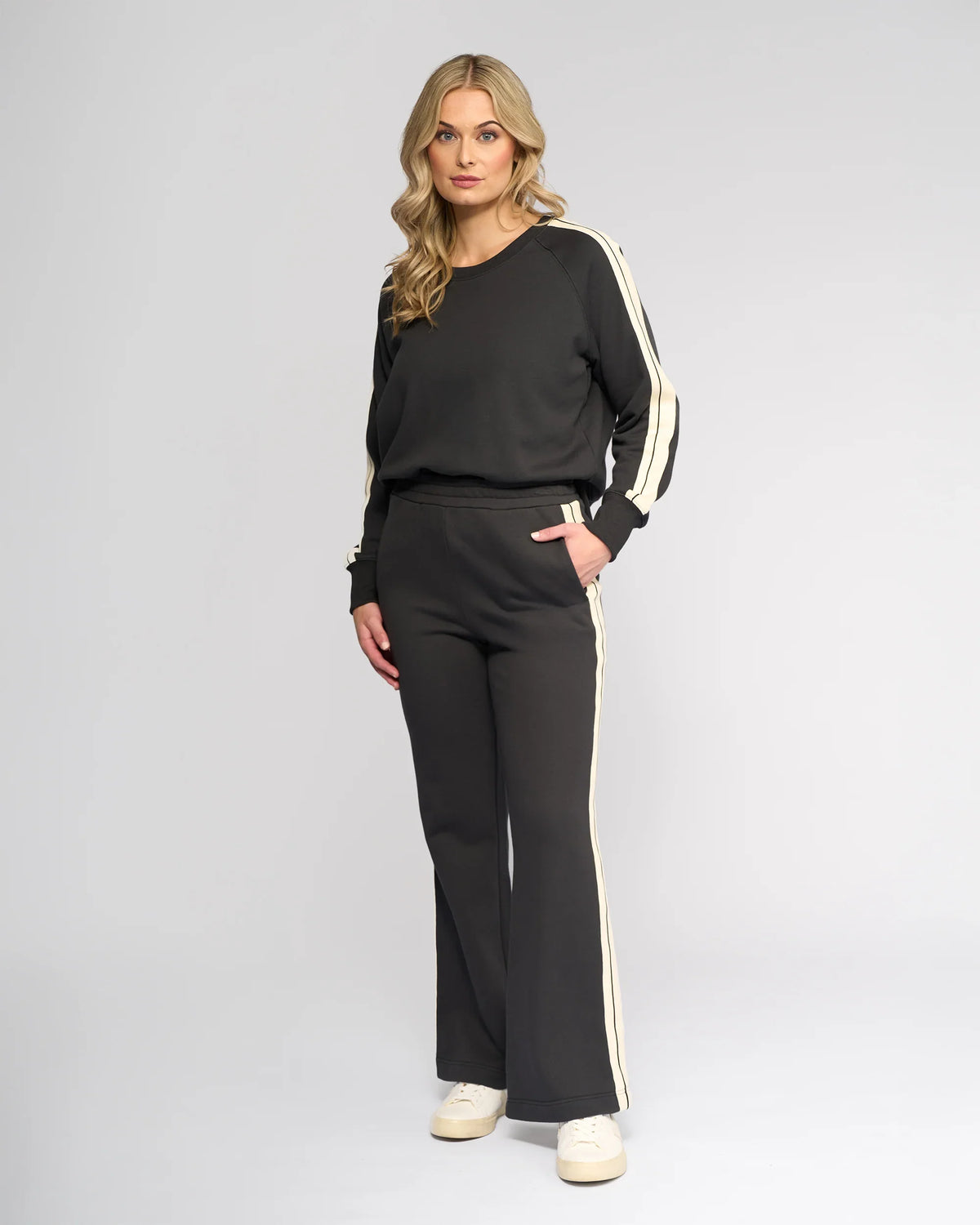 A woman with long blonde hair poses in a studio against a light background, wearing Knewe Label's Rebel Wide Leg - Slate tracksuit and white sneakers, looking directly at the camera.