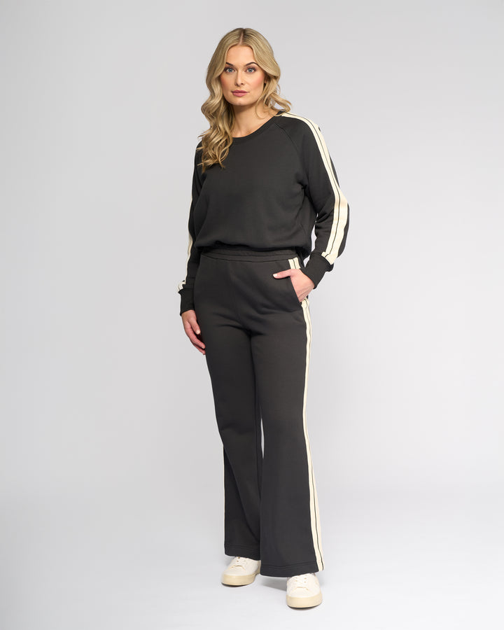 A woman with long blonde hair poses in a studio against a light background, wearing Knewe Label's Rebel Wide Leg - Slate tracksuit and white sneakers, looking directly at the camera.