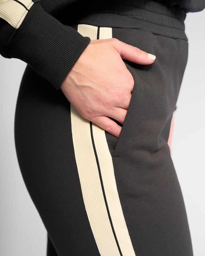 A close-up of someone wearing Knewe Label’s Rebel Wide Leg - Slate pants, featuring cream stripes and a relaxed hand in the side pocket. The cotton fabric contrasts with the plain, light background.
