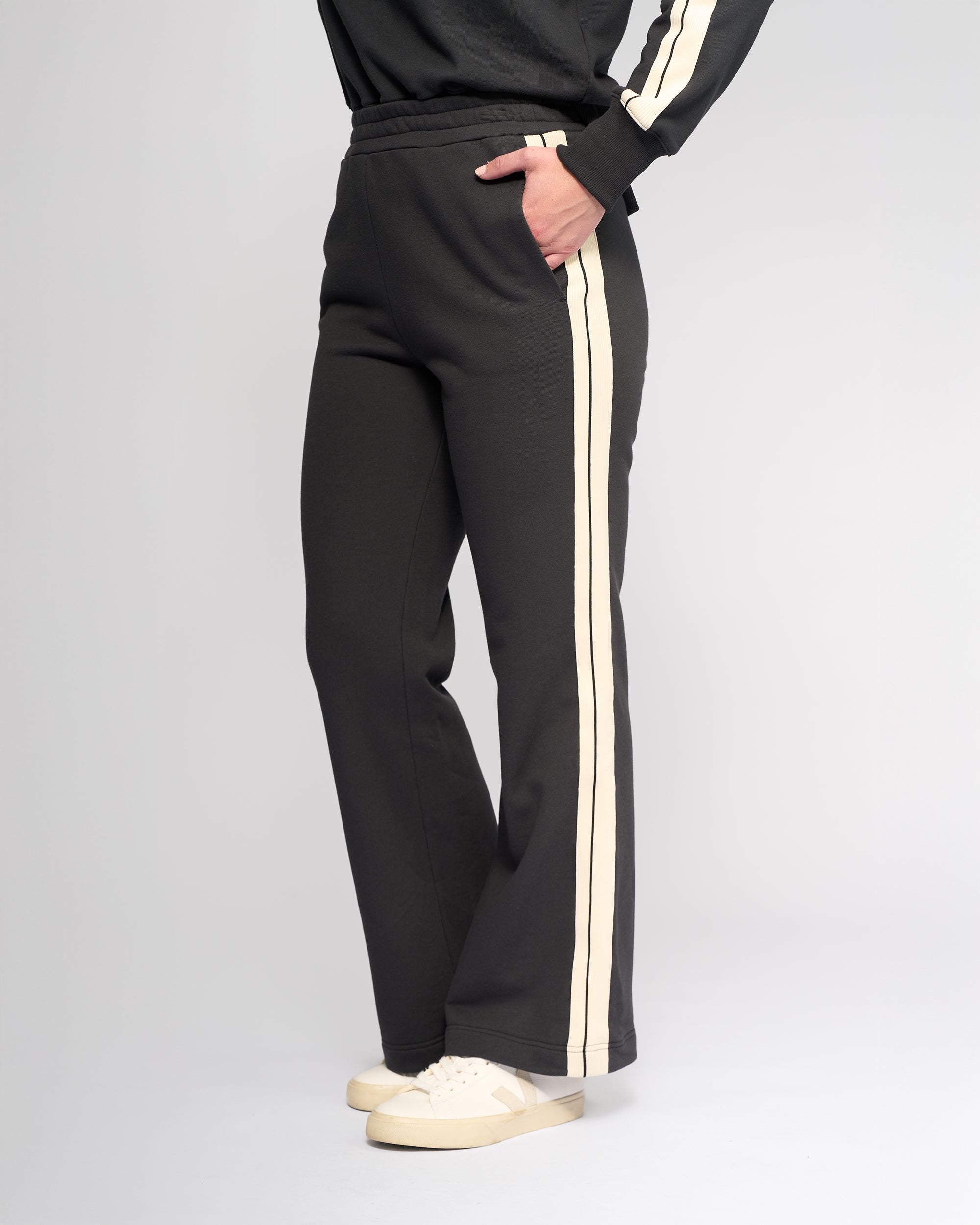 A person wears Knewe Label’s Rebel Wide Leg - Slate sweatpants with white side stripes, paired with a matching black sweatshirt and white sneakers. One hand is in their pocket against a plain light gray background.