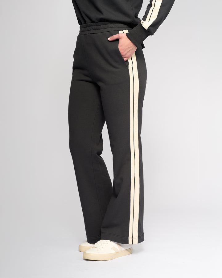 A person wears Knewe Label’s Rebel Wide Leg - Slate sweatpants with white side stripes, paired with a matching black sweatshirt and white sneakers. One hand is in their pocket against a plain light gray background.