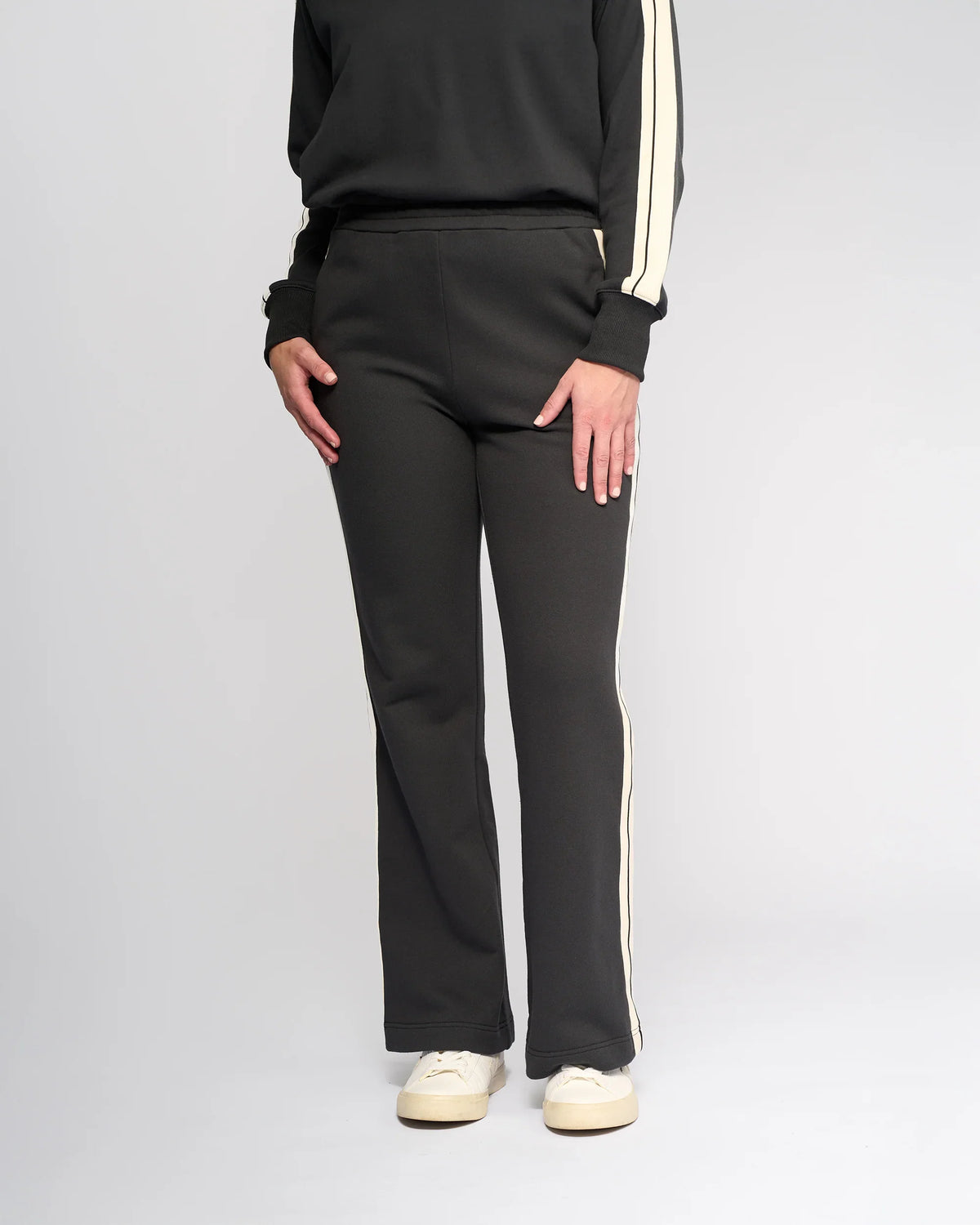 A person wearing Knewe Label’s Rebel Wide Leg - Slate pants and a black top with white stripes, paired with white sneakers, stands against a plain light background.
