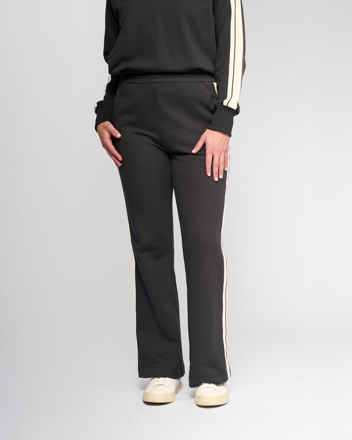 A person wearing Knewe Label’s Rebel Wide Leg - Slate pants and a black top with white stripes, paired with white sneakers, stands against a plain light background.