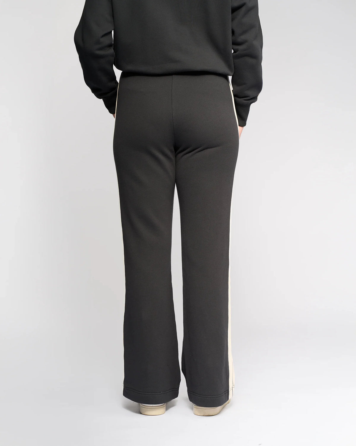 A person faces away from the camera, wearing Knewe Label's Rebel Wide Leg - Slate pants featuring side stripes in soft cotton, paired with light-colored sneakers against a plain white background.