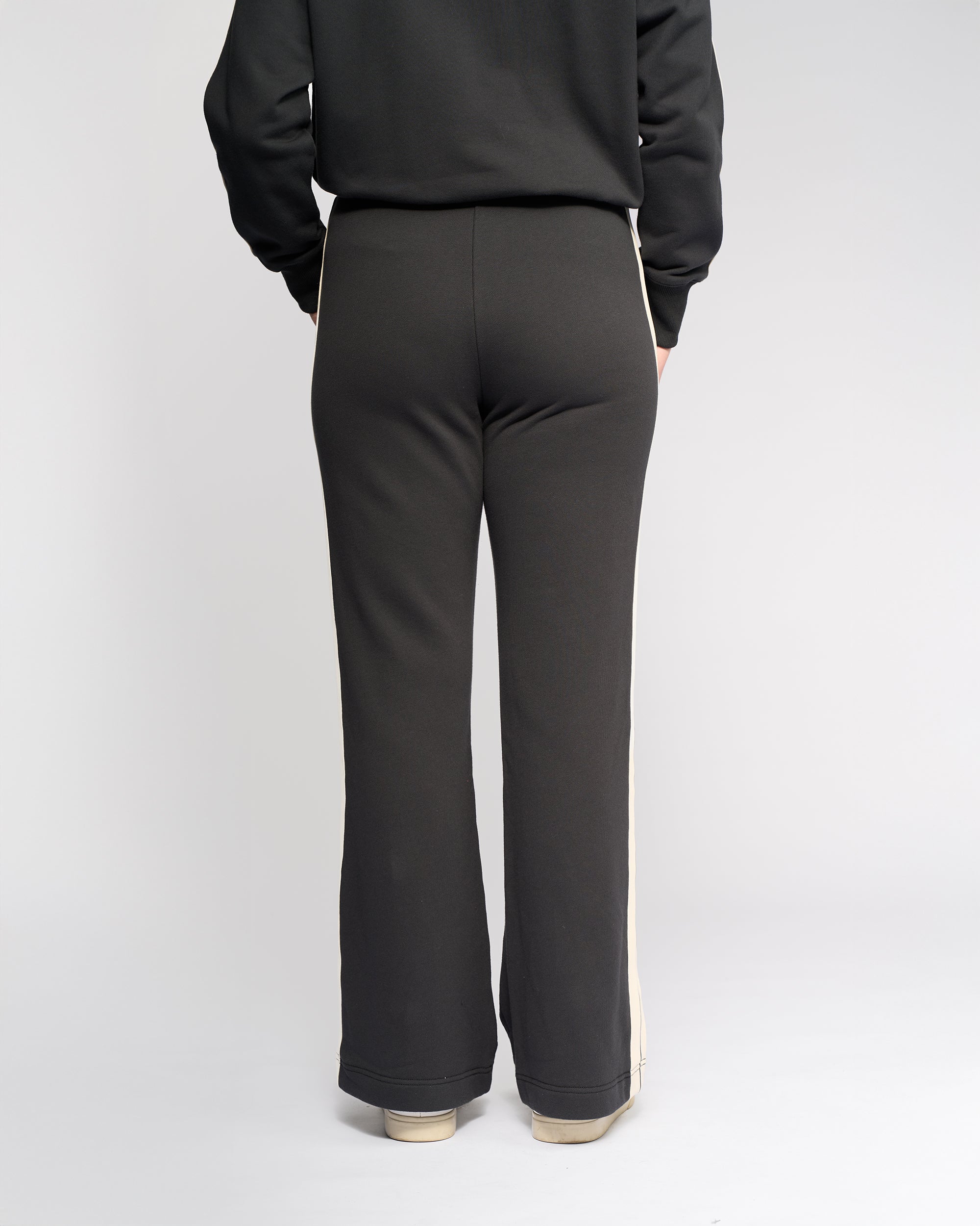 A person faces away from the camera, wearing Knewe Label's Rebel Wide Leg - Slate pants featuring side stripes in soft cotton, paired with light-colored sneakers against a plain white background.