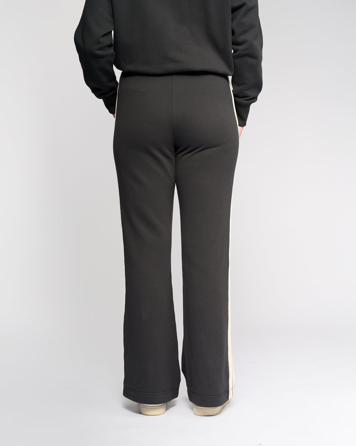 A person faces away from the camera, wearing Knewe Label's Rebel Wide Leg - Slate pants featuring side stripes in soft cotton, paired with light-colored sneakers against a plain white background.