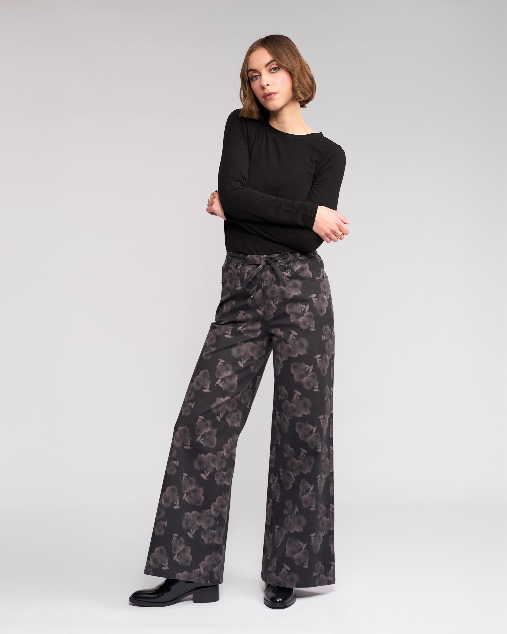 A woman stands with arms crossed, wearing the Knewe Label Goldie Jean - Echo wide-leg pants featuring a subtle abstract echo print, paired with a long-sleeve black top and black shoes, set against a plain light gray background.