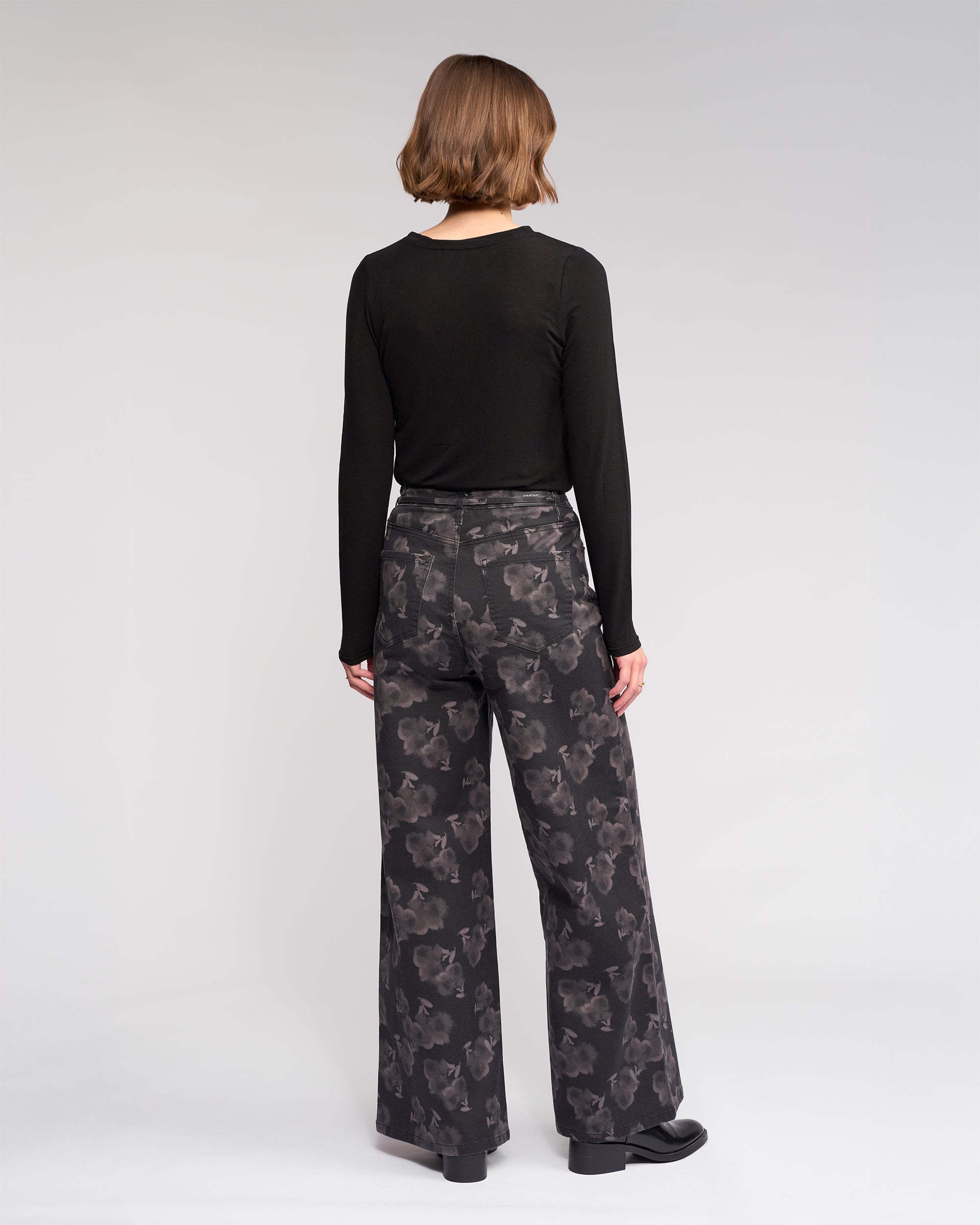 A person with short brown hair faces away, wearing a long-sleeve black top and Knewe Label's Goldie Jean - Echo wide-leg jeans in dark floral denim, against a plain light gray background.